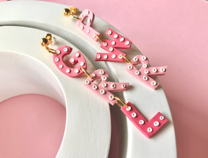 May include: Pink and white letter earrings spelling out "GIRL". Each letter is made of a pink and white ombre material with white dots.