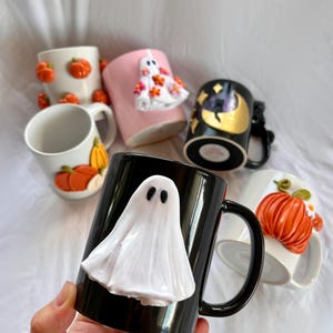 May include: A collection of Halloween themed mugs with various designs. One mug is black with a white ghost, another is pink with a white ghost, another is white with orange pumpkins, another is white with orange pumpkins, and another is black with a gold moon and stars.
