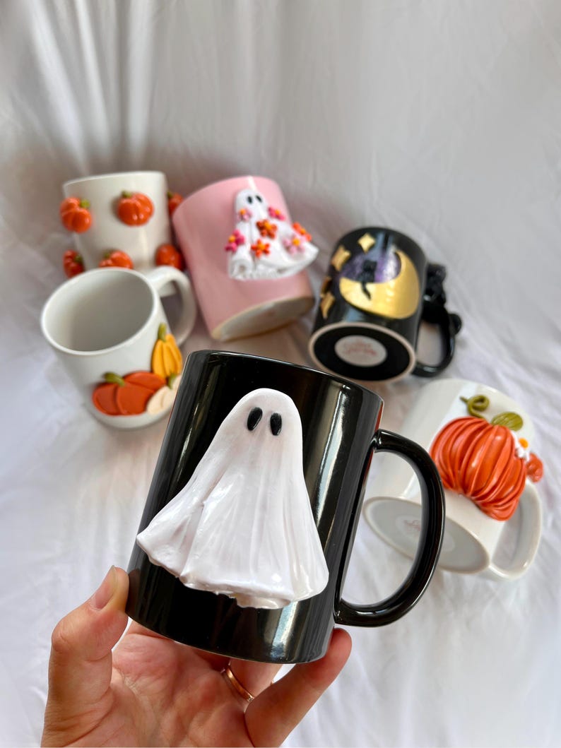 May include: A collection of Halloween themed mugs with various designs. One mug is black with a white ghost, another is pink with a white ghost, another is white with orange pumpkins, another is white with orange pumpkins, and another is black with a gold moon and stars.