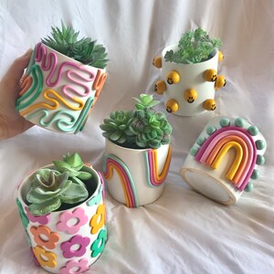May include: Five white ceramic succulent planters with different colourful clay designs. One planter has a rainbow design, one has a smiley face design, one has a flower design, and two have abstract designs.
