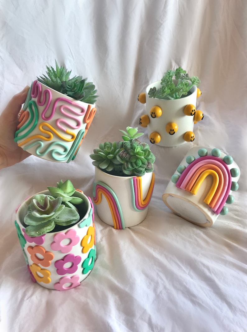 May include: Five white ceramic succulent planters with different colourful clay designs. One planter has a rainbow design, one has a smiley face design, one has a flower design, and two have abstract designs.