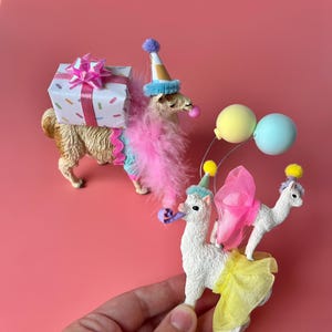 K&ouml;nnte beinhalten: Ein verspieltes Bild mit drei Lama-Figuren. Eines tr&auml;gt ein Geschenk, ein anderes tr&auml;gt einen Partyhut und eine Boa, und das dritte ist mit Luftballons und Partykleidung geschm&uuml;ckt. Der Hintergrund ist einfarbig rosa.