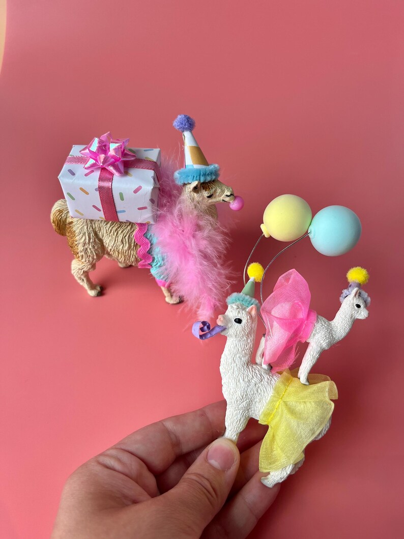 K&ouml;nnte beinhalten: Ein verspieltes Bild mit drei Lama-Figuren. Eines tr&auml;gt ein Geschenk, ein anderes tr&auml;gt einen Partyhut und eine Boa, und das dritte ist mit Luftballons und Partykleidung geschm&uuml;ckt. Der Hintergrund ist einfarbig rosa.