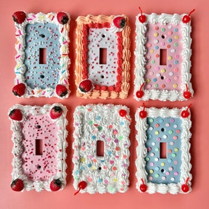 May include: Six light switch covers decorated to look like frosted cakes with colourful sprinkles and cherries. Each cover has a different colour and pattern.
