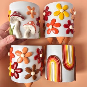 Peut inclure: Quatre pots en céramique blanche avec des motifs différents. Le premier pot a des champignons rouges et blancs et des fleurs orange. Le deuxième pot a des fleurs orange, rouges et jaunes. Le troisième pot a des fleurs orange, rouges, brunes et jaunes. Le quatrième pot a un motif arc-en-ciel en orange, jaune, rouge et blanc.