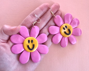 May include: A pair of pink flower-shaped earrings with yellow smiley faces in the center. The earrings are made of polymer clay and have gold hooks.