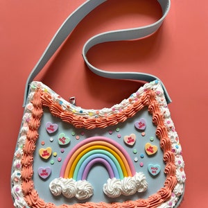 May include: A light blue handbag shaped like a cake with pink frosting, white sprinkles, and a rainbow with clouds. The bag has a light blue strap and features candy hearts with the text "Be Mine" and "Hug Me".