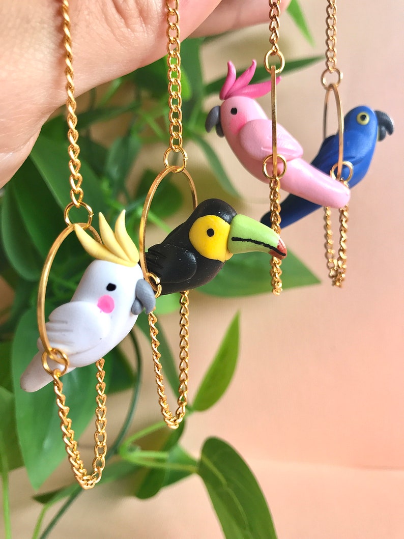 May include: Three colorful bird charms on gold chains. The charms are made of polymer clay and feature a toucan, a cockatoo, and a pink and blue parrot. Each charm is attached to a gold chain with a hoop.