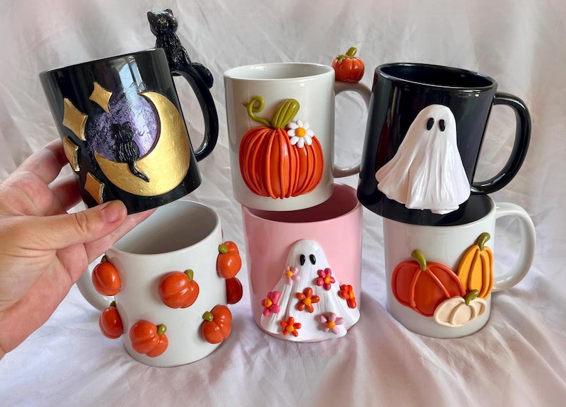May include: Six ceramic mugs with Halloween-themed decorations. The mugs feature pumpkins, ghosts, and a black cat. The mugs are all different colors, including white, black, pink, and gold.