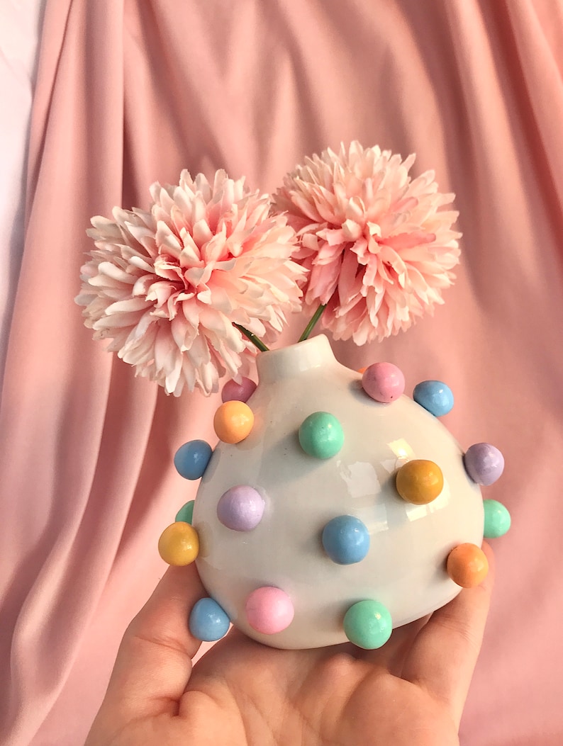 May include: A white ceramic vase with pastel colored polka dots, holding two pink flowers. The vase is round and has a bulbous shape.
