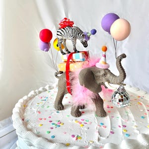May include: A celebratory cake featuring toy animals and balloons. A zebra with a red bow and party hat stands on wrapped gifts. An elephant with a pink boa and party hat holds a disco ball. The white frosted cake is scattered with colourful confetti.