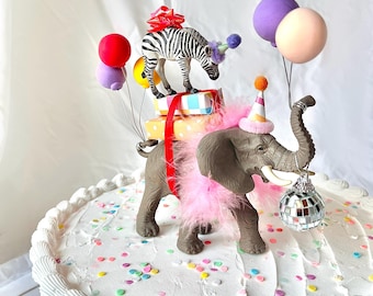 May include: A whimsical cake decorated with toy animals and balloons. A zebra with a red bow and a party hat stands atop wrapped gifts. An elephant with a pink boa and party hat holds a disco ball. Colorful confetti dots the white frosted cake.