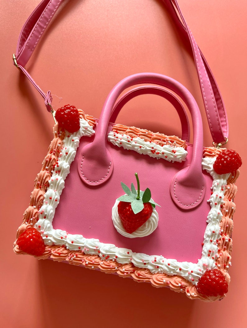 May include: A pink faux leather handbag shaped like a cake with white frosting and red and pink sprinkles. The bag has a strawberry on top and a detachable strap.