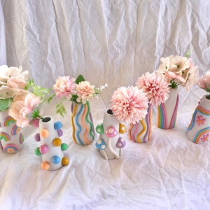 May include: Seven small ceramic vases with different pastel colored designs. The vases are filled with pink flowers and are arranged on a white surface.