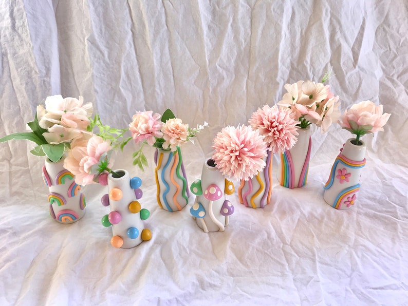 May include: Seven small ceramic vases with different pastel colored designs. The vases are filled with pink flowers and are arranged on a white surface.