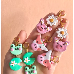 Pu&ograve; includere: Un set di sei paia di orecchini pendenti con vari disegni di animali e fiori. Gli orecchini sono realizzati in pasta polimerica e presentano una rana verde, un maiale rosa, una mucca con una corona di fragole, una margherita e una ninfea verde.