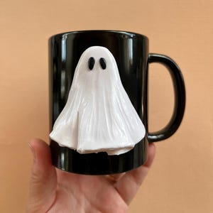 May include: Black ceramic mug with a white ghost sculpted on the front. The ghost has two black eyes.