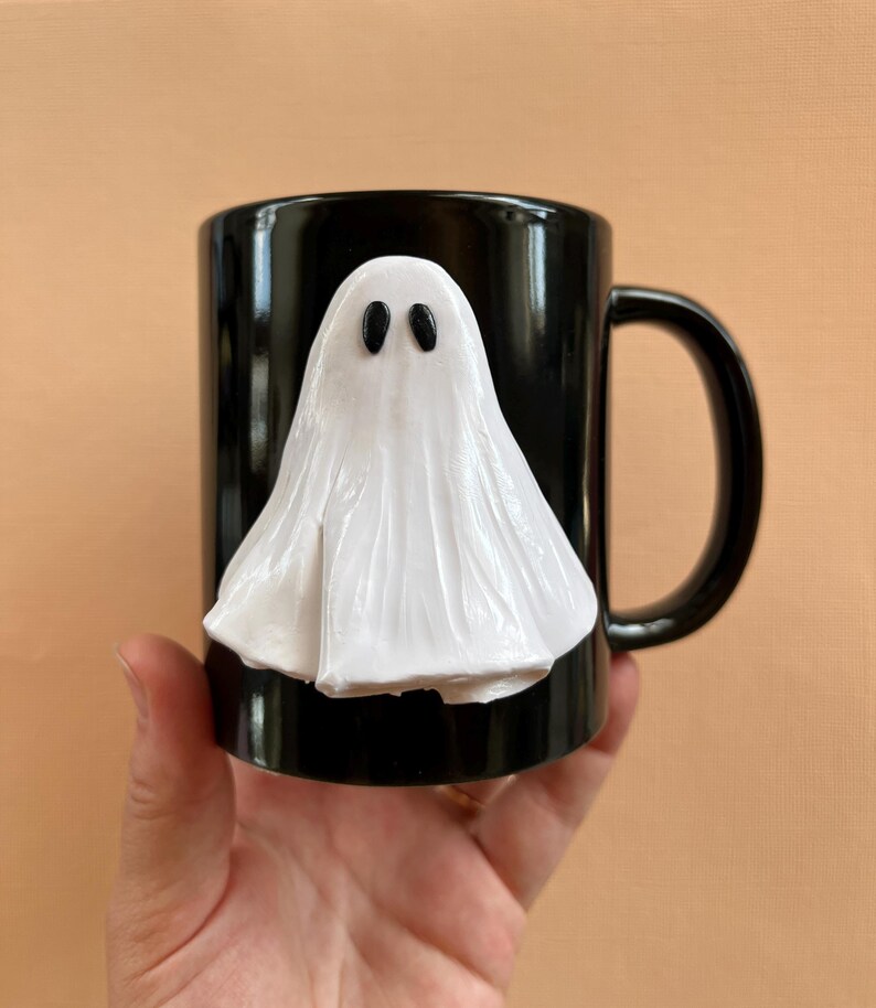May include: Black ceramic mug with a white ghost sculpted on the front. The ghost has two black eyes.
