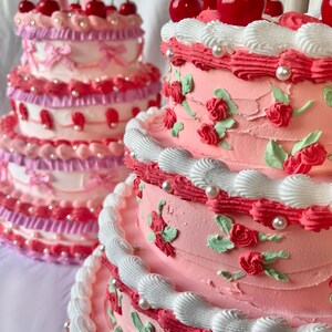 May include: Two tiered cakes decorated with pink and white frosting, red cherries, and pearl accents. The cake on the left has a pink and white layered design, while the cake on the right has a more traditional white frosting with red roses and green leaves.