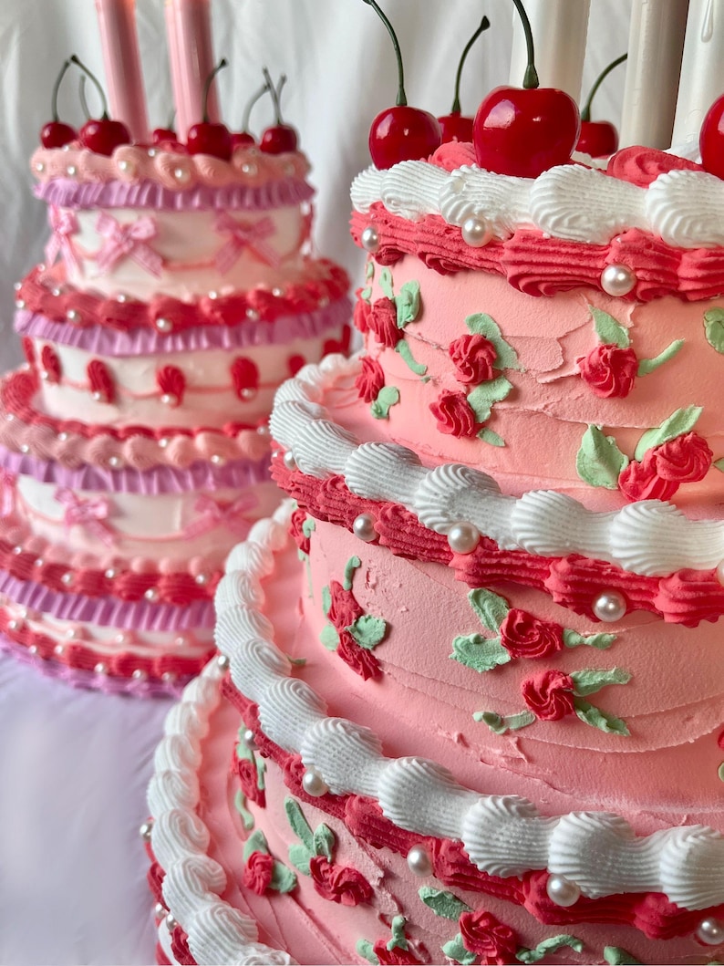 May include: Two tiered cakes decorated with pink and white frosting, red cherries, and pearl accents. The cake on the left has a pink and white layered design, while the cake on the right has a more traditional white frosting with red roses and green leaves.