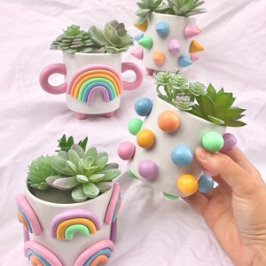 May include: Four small white ceramic planters with different designs. The planters are decorated with pastel-colored rainbows, polka dots, and spikes. Each planter contains a small green succulent plant.