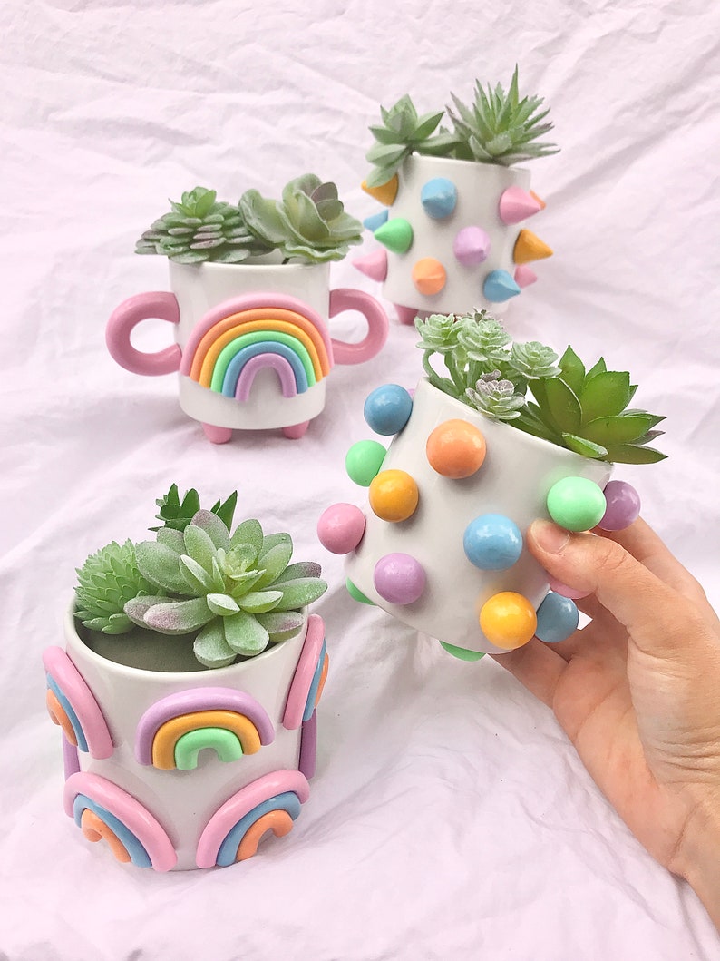 May include: Four small white ceramic planters with different designs. The planters are decorated with pastel-colored rainbows, polka dots, and spikes. Each planter contains a small green succulent plant.