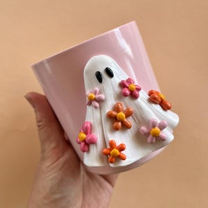 May include: A pink ceramic planter with a white ghost figurine decorated with pink, orange, and yellow flowers.