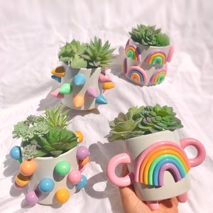May include: Four colorful ceramic planters with succulents. The planters are decorated with rainbow designs, spikes, and colorful dots.