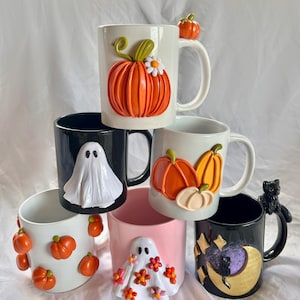 May include: Six ceramic mugs with Halloween-themed decorations. The mugs are stacked in a pyramid shape. The top mug is white with an orange pumpkin and a small white flower. The second mug is black with a white ghost. The third mug is white with two orange pumpkins. The fourth mug is pink with a white ghost and orange and pink flowers. The fifth mug is black with a gold moon and a black cat. The sixth mug is white with orange pumpkins.