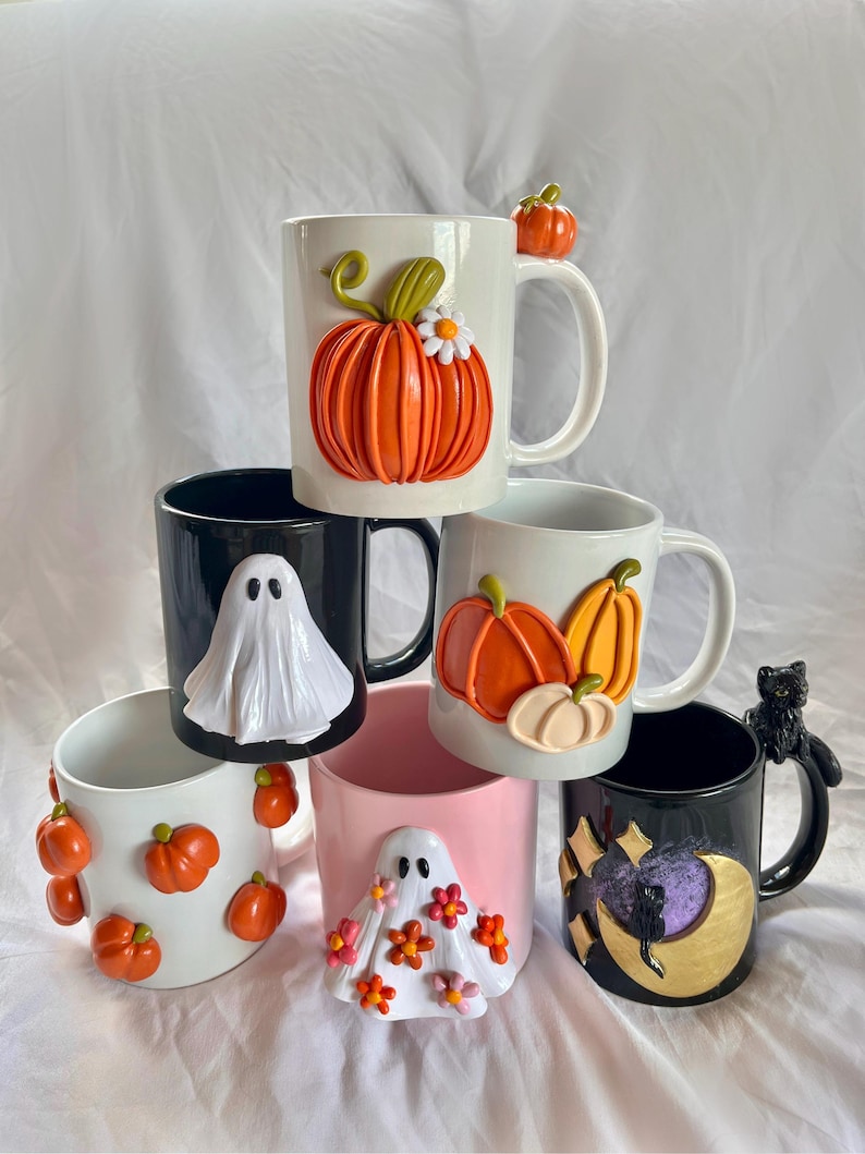 May include: Six ceramic mugs with Halloween-themed decorations. The mugs are stacked in a pyramid shape. The top mug is white with an orange pumpkin and a small white flower. The second mug is black with a white ghost. The third mug is white with two orange pumpkins. The fourth mug is pink with a white ghost and orange and pink flowers. The fifth mug is black with a gold moon and a black cat. The sixth mug is white with orange pumpkins.