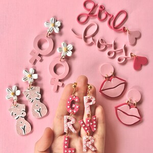 May include: Pink and white polymer clay earrings with various designs, including a female symbol, a daisy, a pair of breasts, and the letters "GIRL" and "BOY".