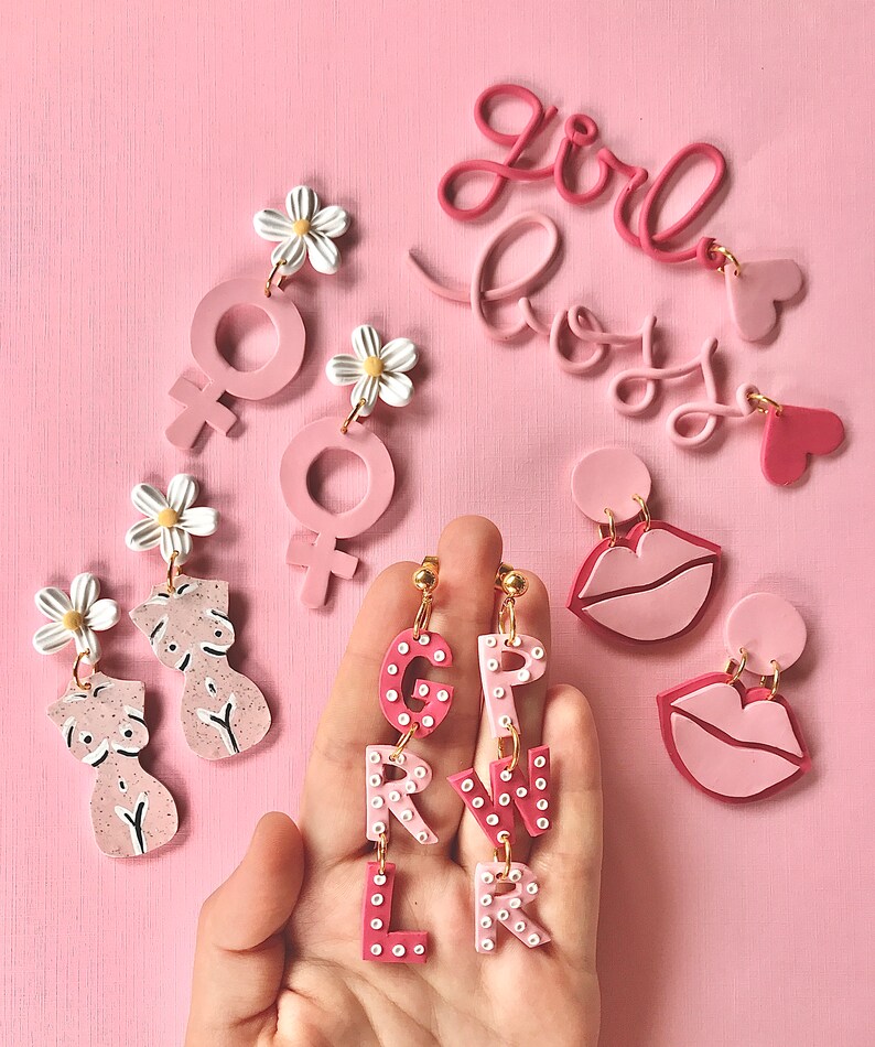 May include: Pink and white polymer clay earrings with various designs, including a female symbol, a daisy, a pair of breasts, and the letters "GIRL" and "BOY".