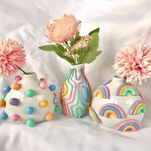 May include: Three ceramic vases with different decorative designs. The vase on the left is white with colorful polka dots. The middle vase is white with colorful wavy lines. The vase on the right is white with colorful rainbow designs.
