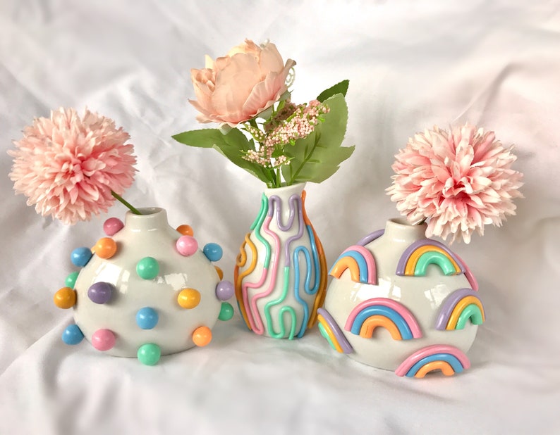 May include: Three ceramic vases with different decorative designs. The vase on the left is white with colorful polka dots. The middle vase is white with colorful wavy lines. The vase on the right is white with colorful rainbow designs.