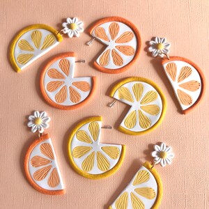 May include: A pair of earrings featuring a half-circle lemon slice with a white daisy charm. The lemon slices are made of polymer clay and have a yellow and orange color scheme.