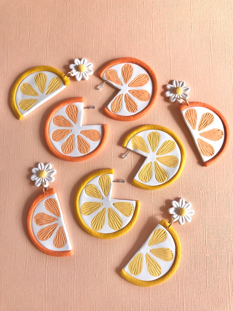 May include: A pair of earrings featuring a half-circle lemon slice with a white daisy charm. The lemon slices are made of polymer clay and have a yellow and orange color scheme.
