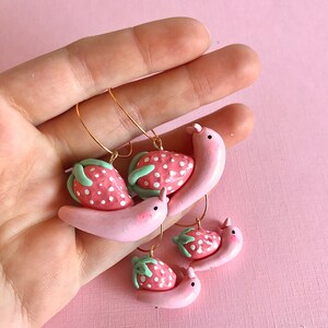 Pu&ograve; includere: Tre paia di orecchini in pasta polimerica rosa e verde a forma di lumache che trasportano fragole. Gli orecchini sono su uno sfondo rosa.
