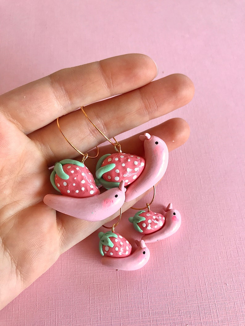 Pu&ograve; includere: Tre paia di orecchini in pasta polimerica rosa e verde a forma di lumache che trasportano fragole. Gli orecchini sono su uno sfondo rosa.