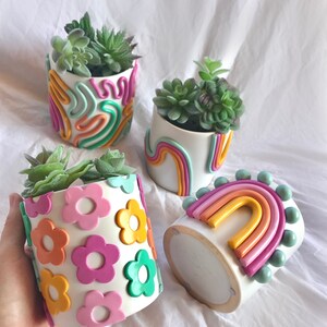 May include: Four white ceramic plant pots with different colourful clay designs. One pot has a rainbow design, another has a flower design, and two have abstract designs. Each pot contains a small succulent plant.