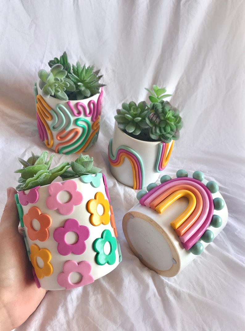 May include: Four white ceramic plant pots with different colourful clay designs. One pot has a rainbow design, another has a flower design, and two have abstract designs. Each pot contains a small succulent plant.