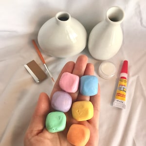 May include: Six small, colorful, square-shaped clay pieces in a hand. The clay pieces are pink, light blue, lavender, yellow, green, and orange. There are two white ceramic vases, a small jar of white glue, a tube of super glue, a paintbrush, and a piece of sandpaper in the background.