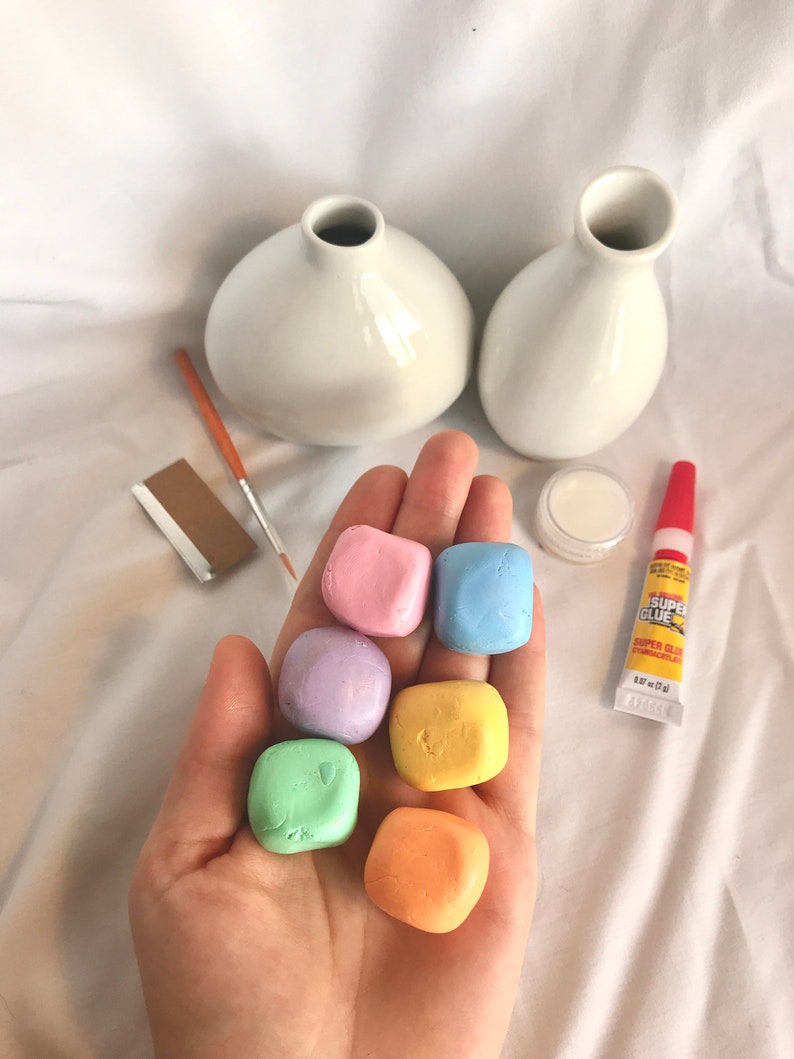 May include: Six small, colorful, square-shaped clay pieces in a hand. The clay pieces are pink, light blue, lavender, yellow, green, and orange. There are two white ceramic vases, a small jar of white glue, a tube of super glue, a paintbrush, and a piece of sandpaper in the background.