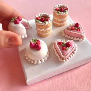 May include: Six miniature cakes decorated with strawberries and white frosting. The cakes are arranged on a white display stand. The cakes are made of polymer clay and are perfect for jewelry making or other craft projects.