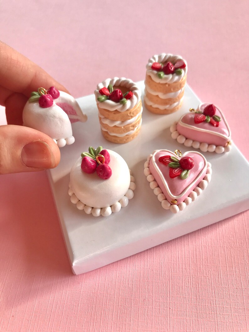 May include: Six miniature cakes decorated with strawberries and white frosting. The cakes are arranged on a white display stand. The cakes are made of polymer clay and are perfect for jewelry making or other craft projects.