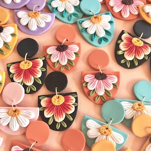 May include: Assortment of handmade polymer clay earrings featuring floral designs. The earrings are in various colors, including black, white, orange, pink, and green. Each earring features a unique flower design, with some featuring a single flower and others featuring multiple flowers.
