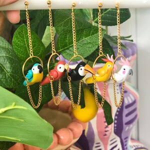 May include: Five pairs of dangle earrings featuring different colorful bird charms. Each bird is perched on a gold hoop and hangs from a gold chain.