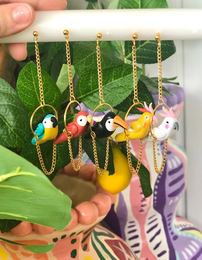 May include: Five pairs of dangle earrings featuring different colorful bird charms. Each bird is perched on a gold hoop and hangs from a gold chain.