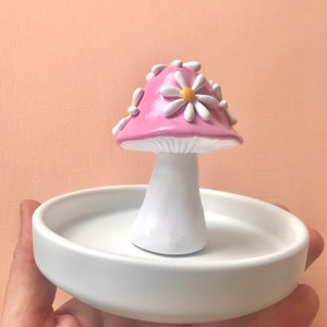 Puede incluir: Un porta anillos de hongo rosa con flores de margarita blancas en la parte superior. El hongo est&aacute; sentado en un plato de cer&aacute;mica blanco.