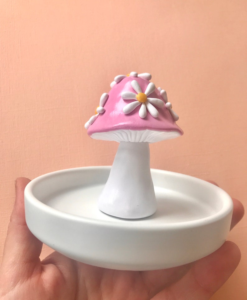 Puede incluir: Un porta anillos de hongo rosa con flores de margarita blancas en la parte superior. El hongo est&aacute; sentado en un plato de cer&aacute;mica blanco.