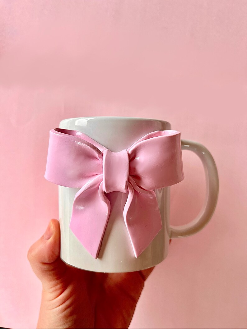 Pu&ograve; includere: Una tazza in ceramica bianca con un grande fiocco rosa attaccato alla parte anteriore. La tazza &egrave; tenuta in una mano.
